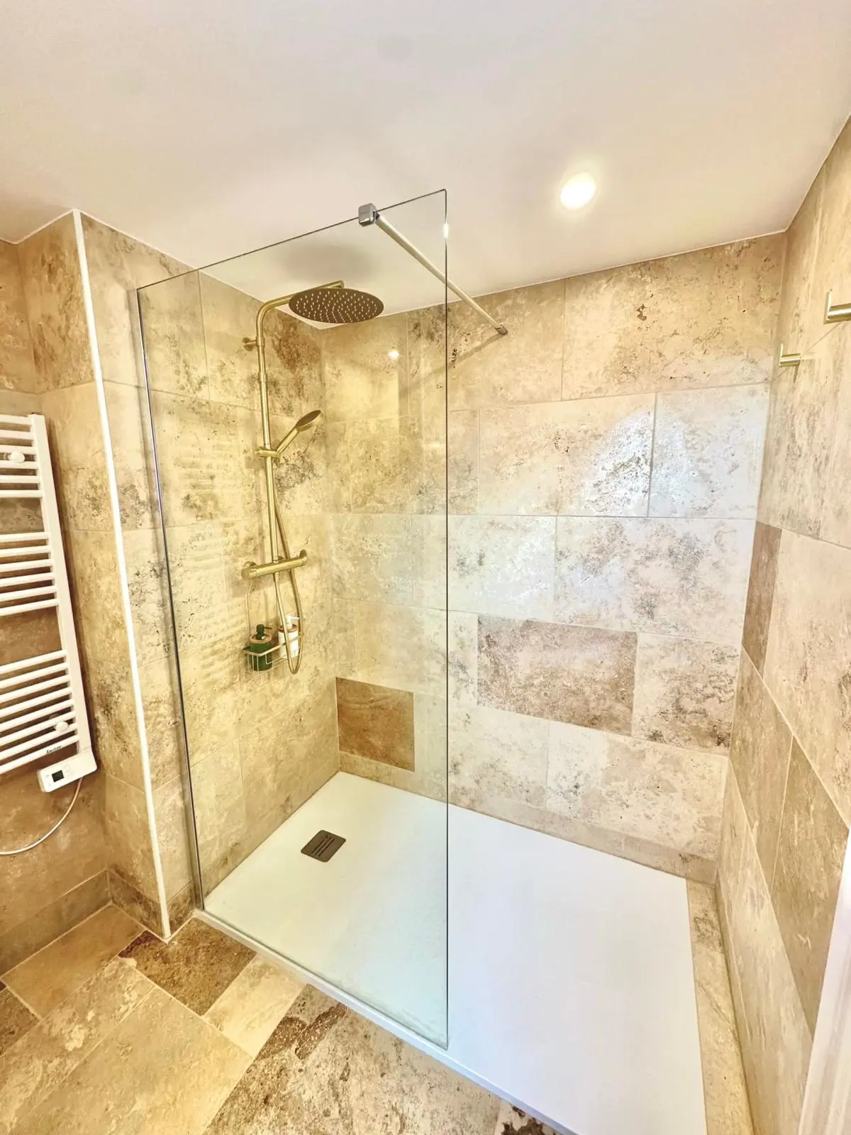 Après rénovation — douche à l'italienne en travertin avec receveur extra-plat blanc, paroi fixe et colonne de douche dorée