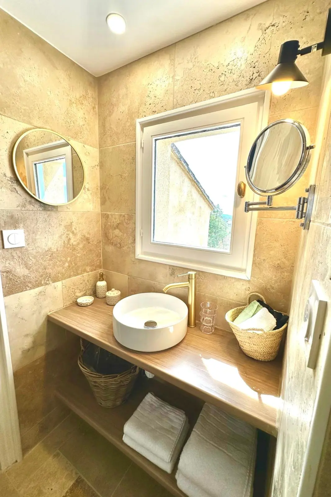 Après rénovation — vasque à poser sur plan bois, robinet laiton brossé et murs en travertin sous la fenêtre