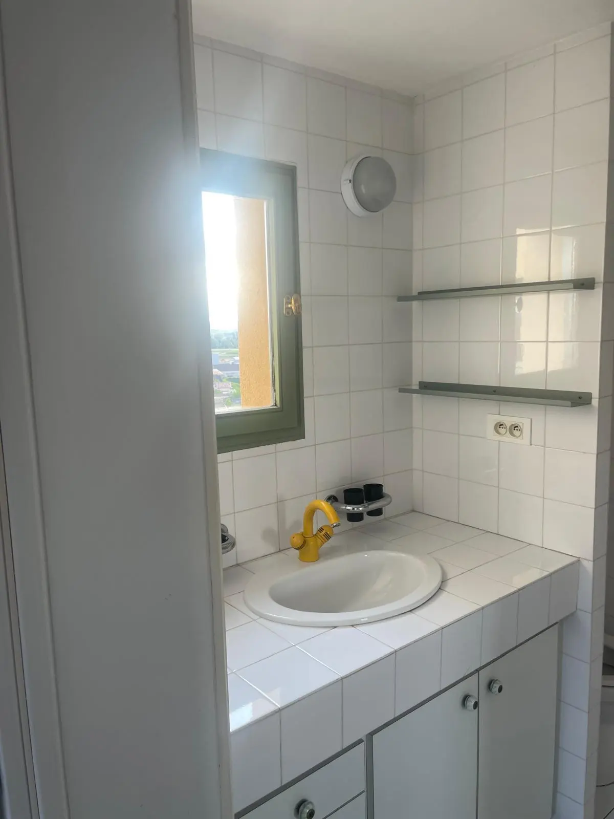 Avant rénovation — lavabo encastré dans un plan carrelé blanc avec robinet jaune et hublot d'éclairage