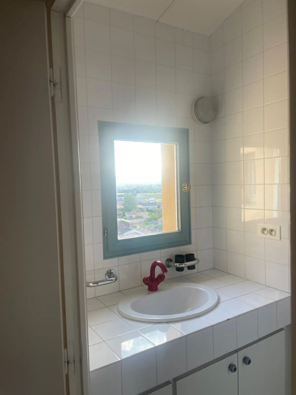 Avant rénovation — lavabo encastré dans un plan carrelé blanc avec robinet rouge sous la fenêtre